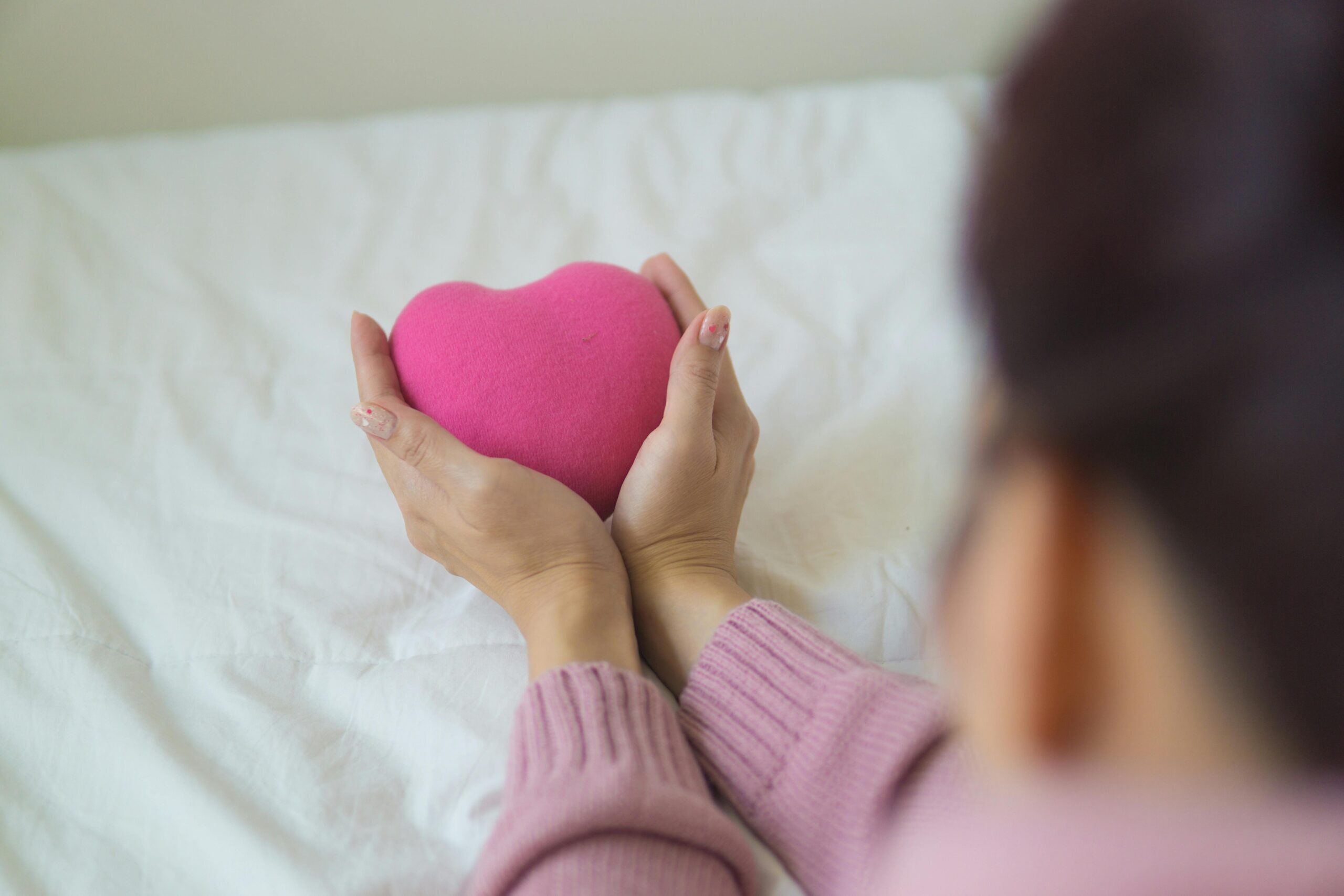 bedsheets with woman in light pink jumper and brown hair holding arms out and hands cradling a pink felt heart