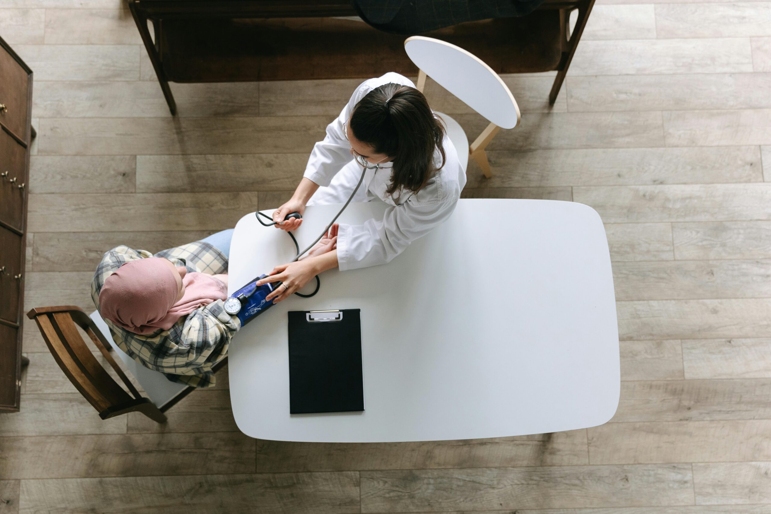 Overhead view wooden floor white table woman in flannel shirt and pink headscarf sits while a woman in a white doctors coat and brown hair reaches out to check her blood pressure with a sleeve