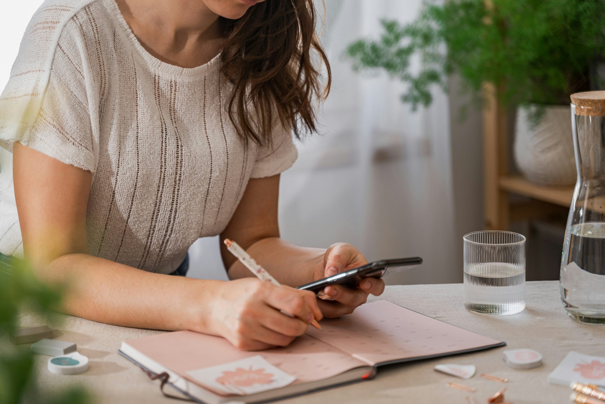 woman visible from neck down in beige stripe knit top sitting at table with water and water jug leaning over salmon pink weekly planner holding her phone in left hand and pen in other with sticky notes surrounding and one on page