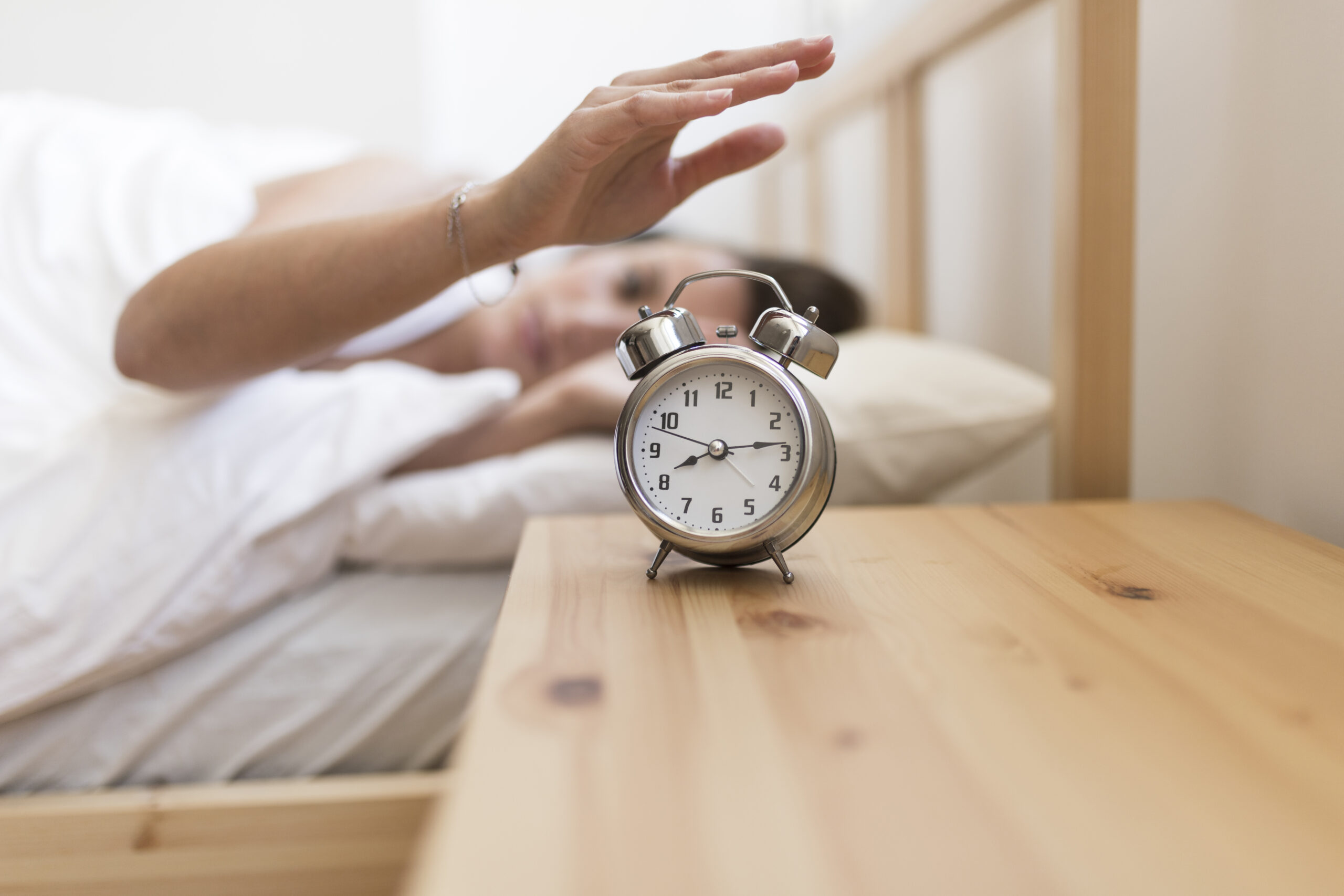 woman in ben wooden bedframe with white sheets on bed reaching out to matching wooden side table to turn off silver alarm clock set at time 8.13
