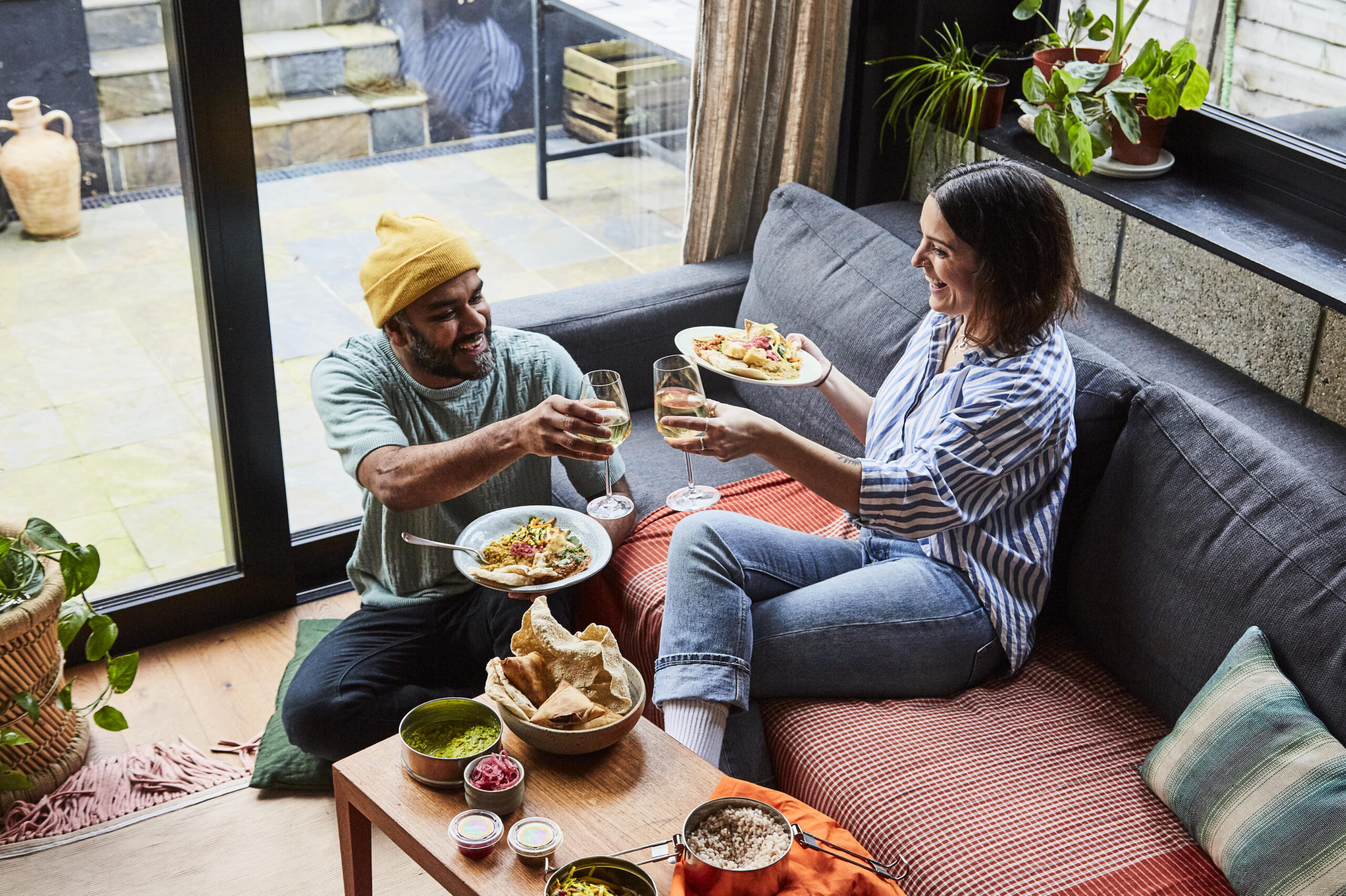 man and woman sitting on sofa sharing a meal and cheersing wines