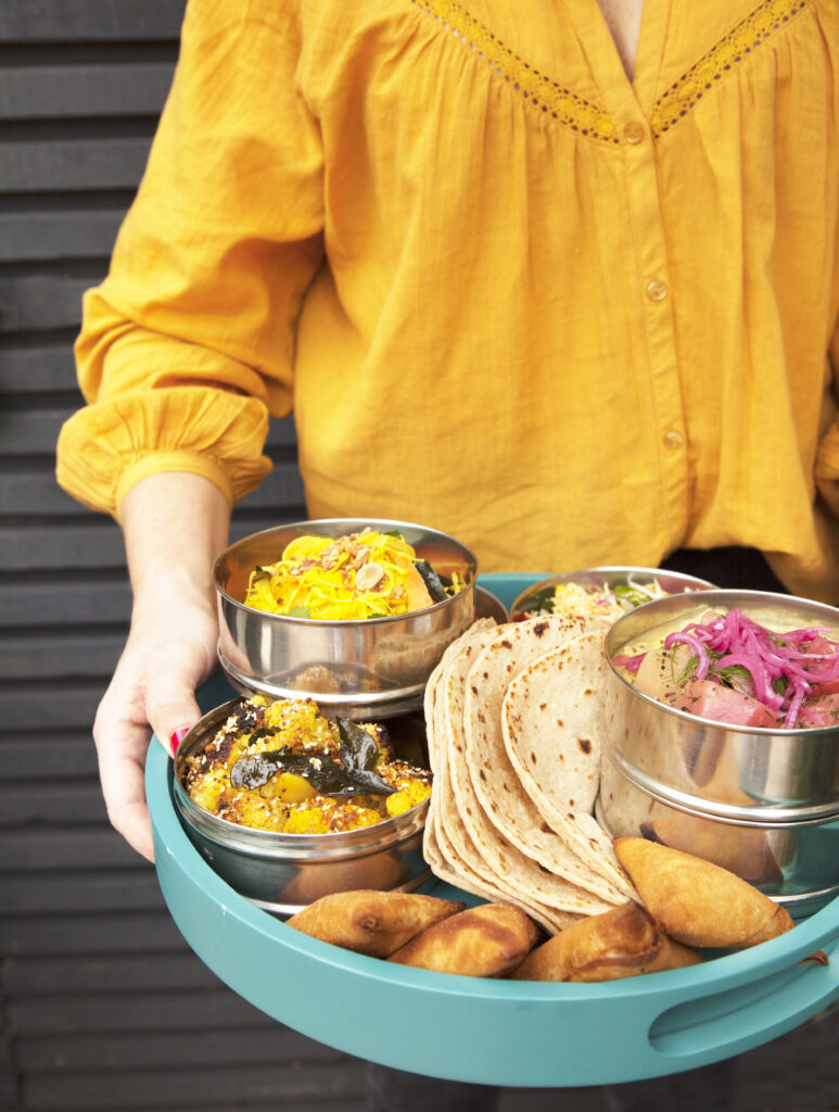 woman in yellow long sleeve top holding out a tray of curry and bread and samosas in tiffin tins