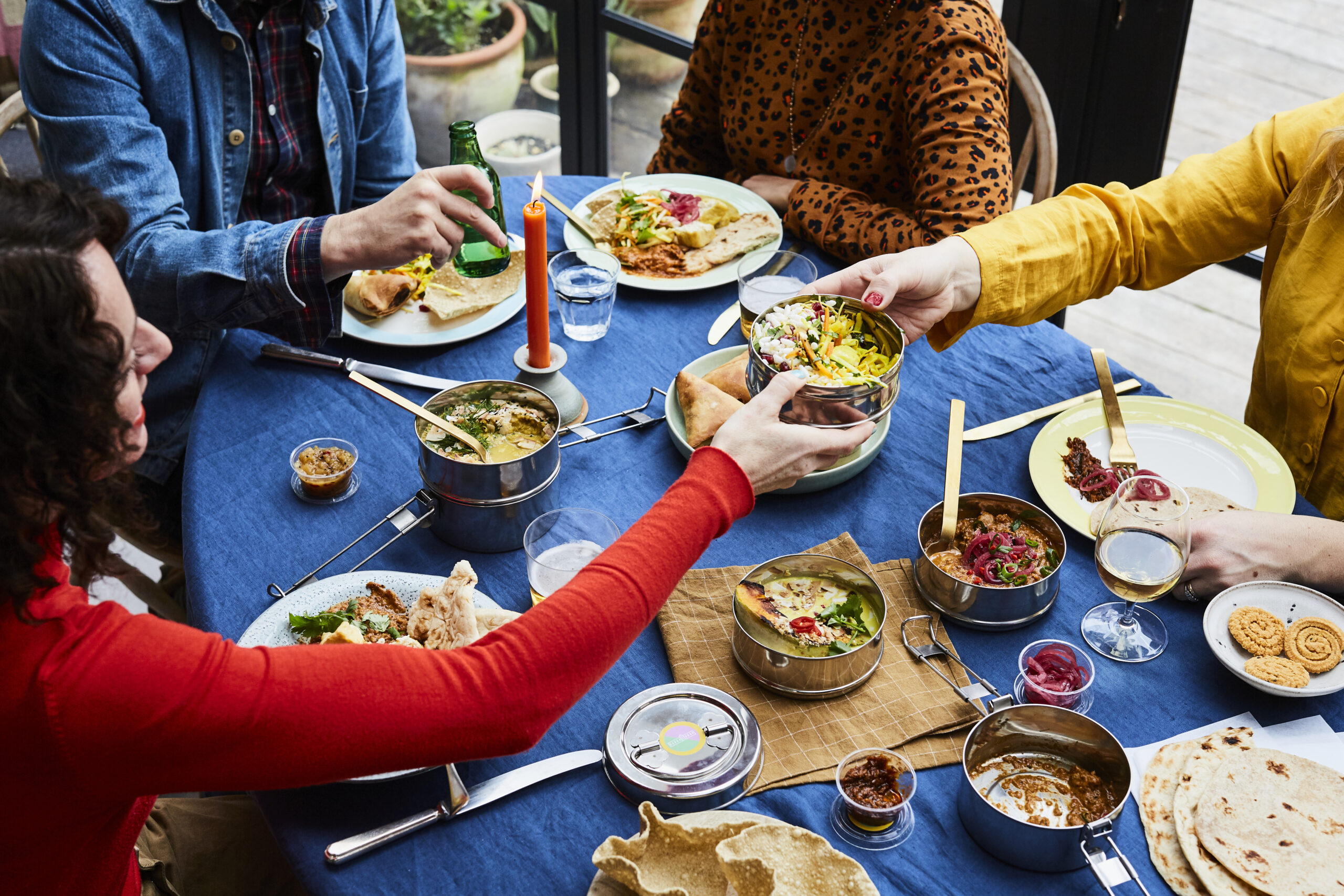blue table with dinner laid out in tiffin tins woman in red top is handing a tiffin tin over the table with plates with food on