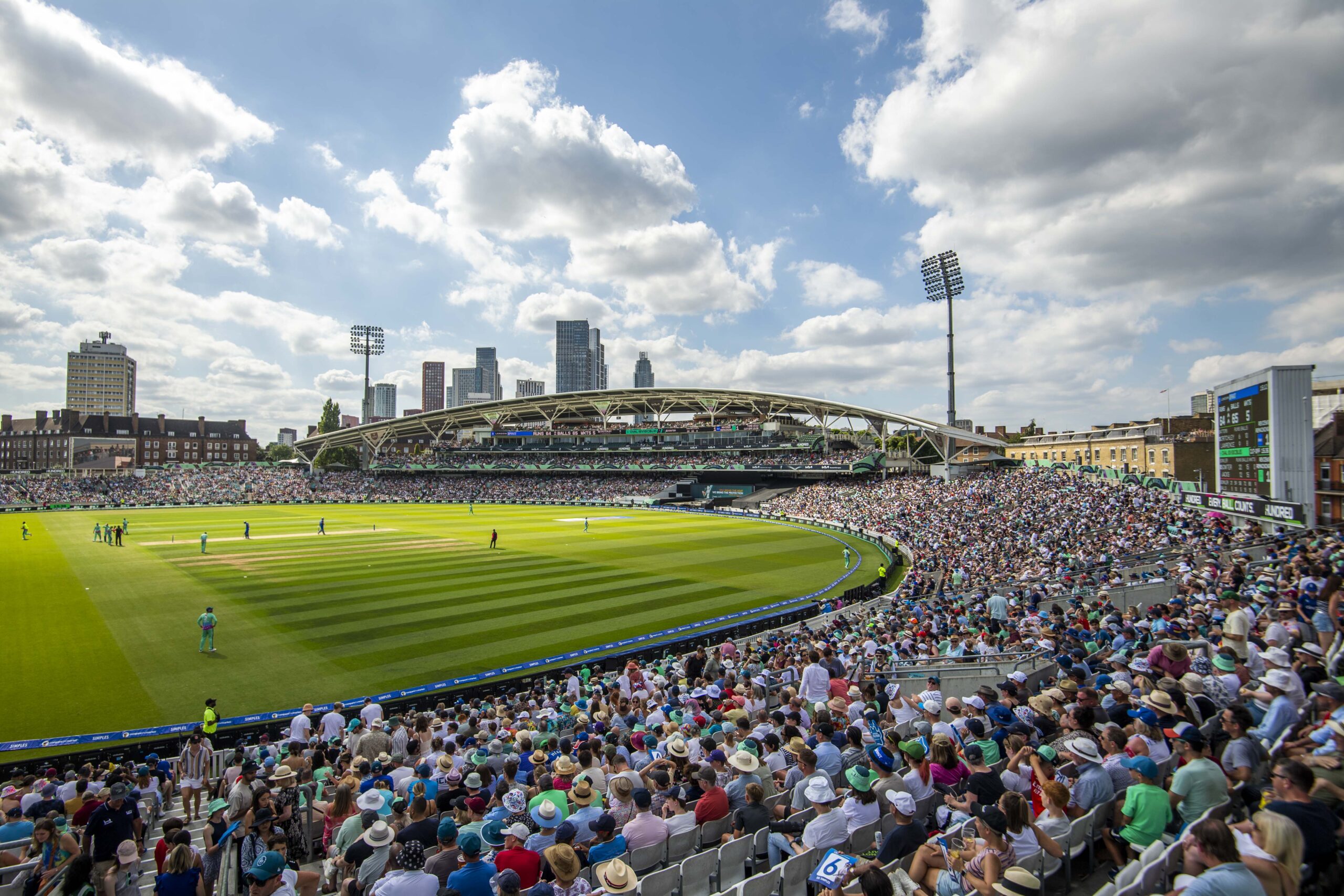 side view of a cricket stadium from the crowd with a green pitch and lots of people in the stands and a sky with clouds