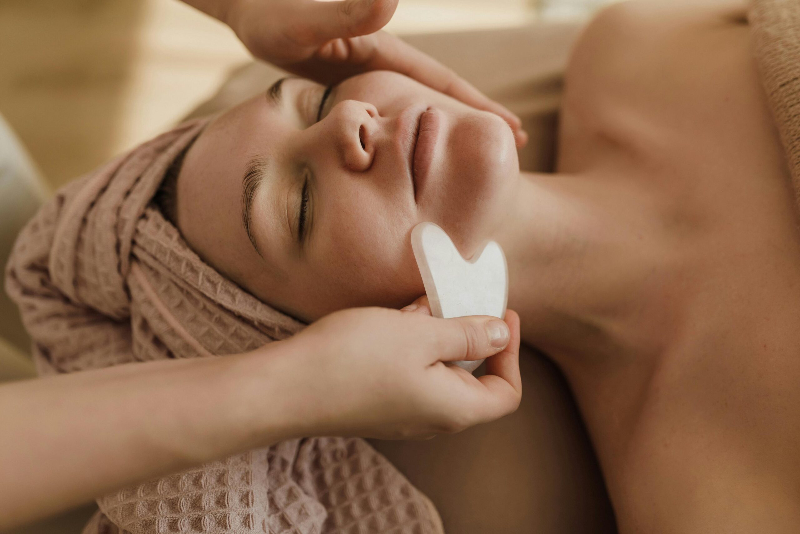 Woman lying back on treatment table with pink waffle towel on head receiving lymphatic massage with a guasha 
