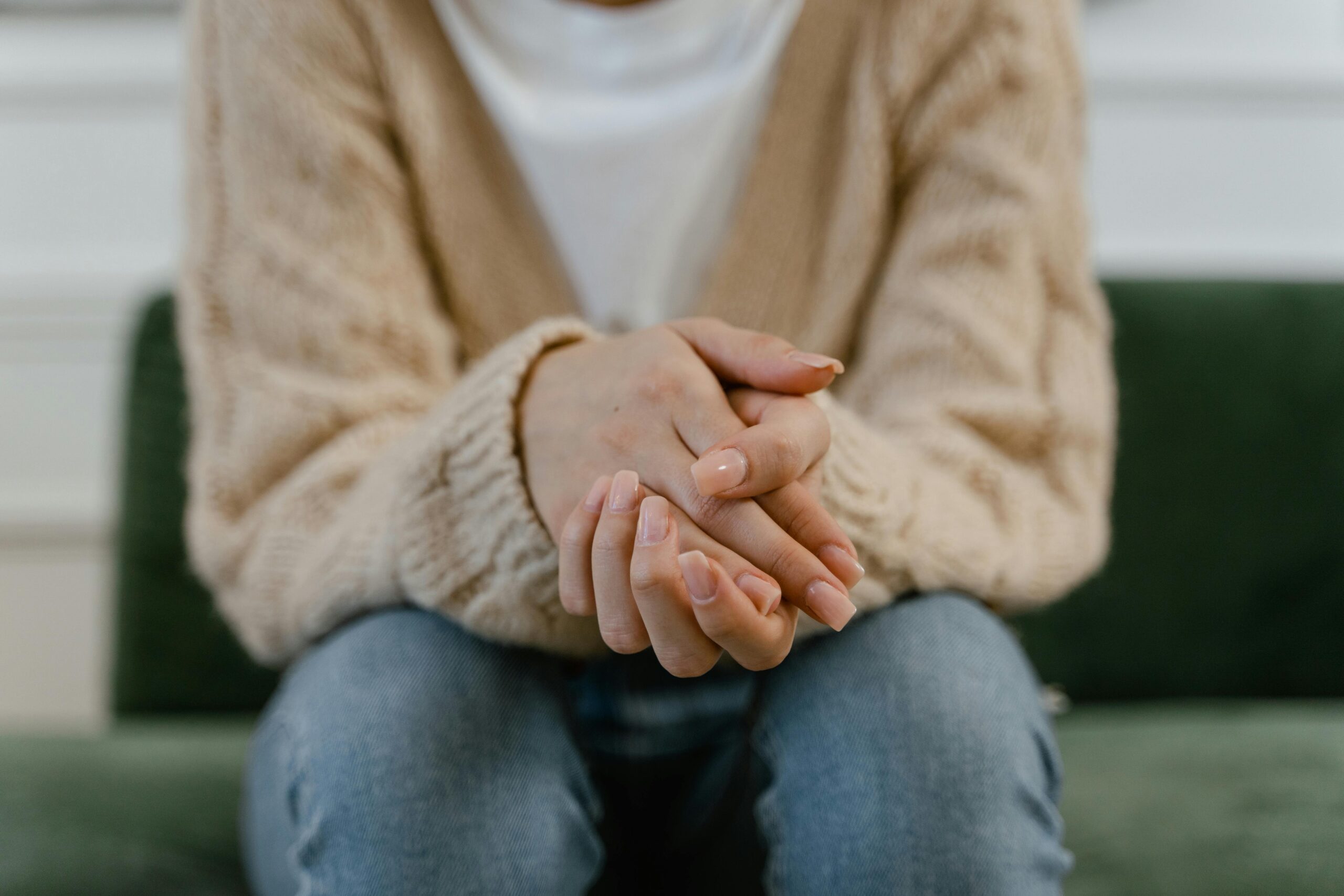 woman in knit jumper and jeans sitting on green sofa with hands clasped in front of her