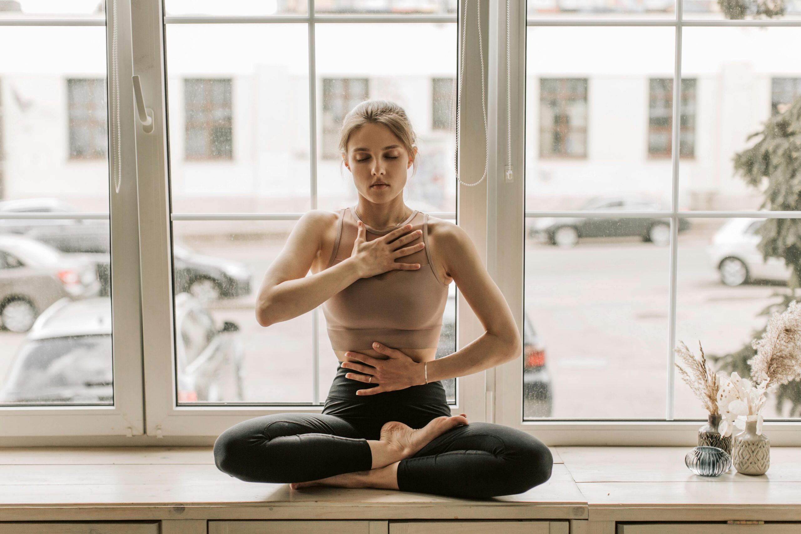 woman sits indoors on a ledge by a window with her legs crossed and her hands over her body one on her chest and the other on her middle torso
