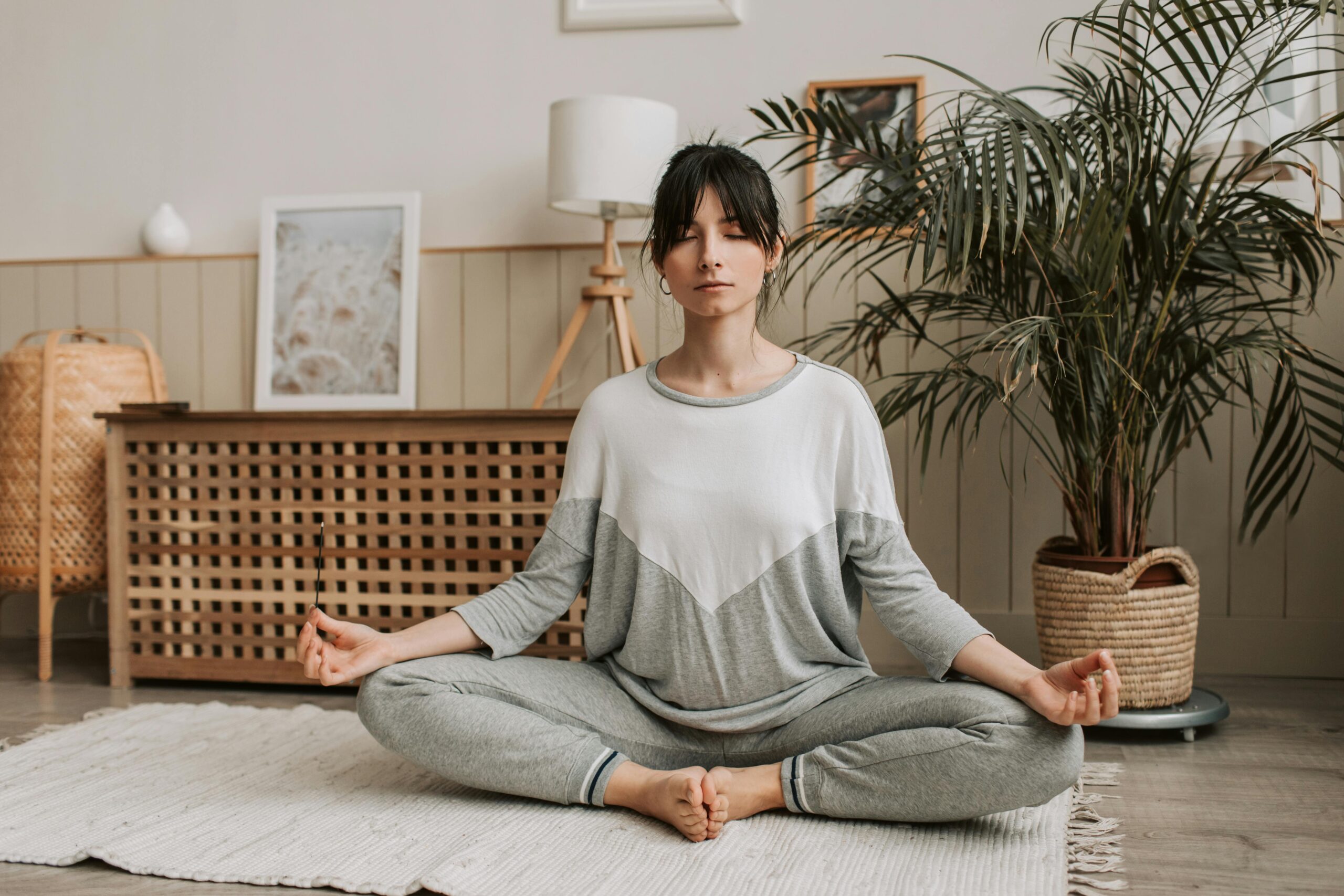 Woman sitting on the ground in room with blanket on floor and plant behind wearing grey sweatpants in yoga poses with arms either side in meditation pose