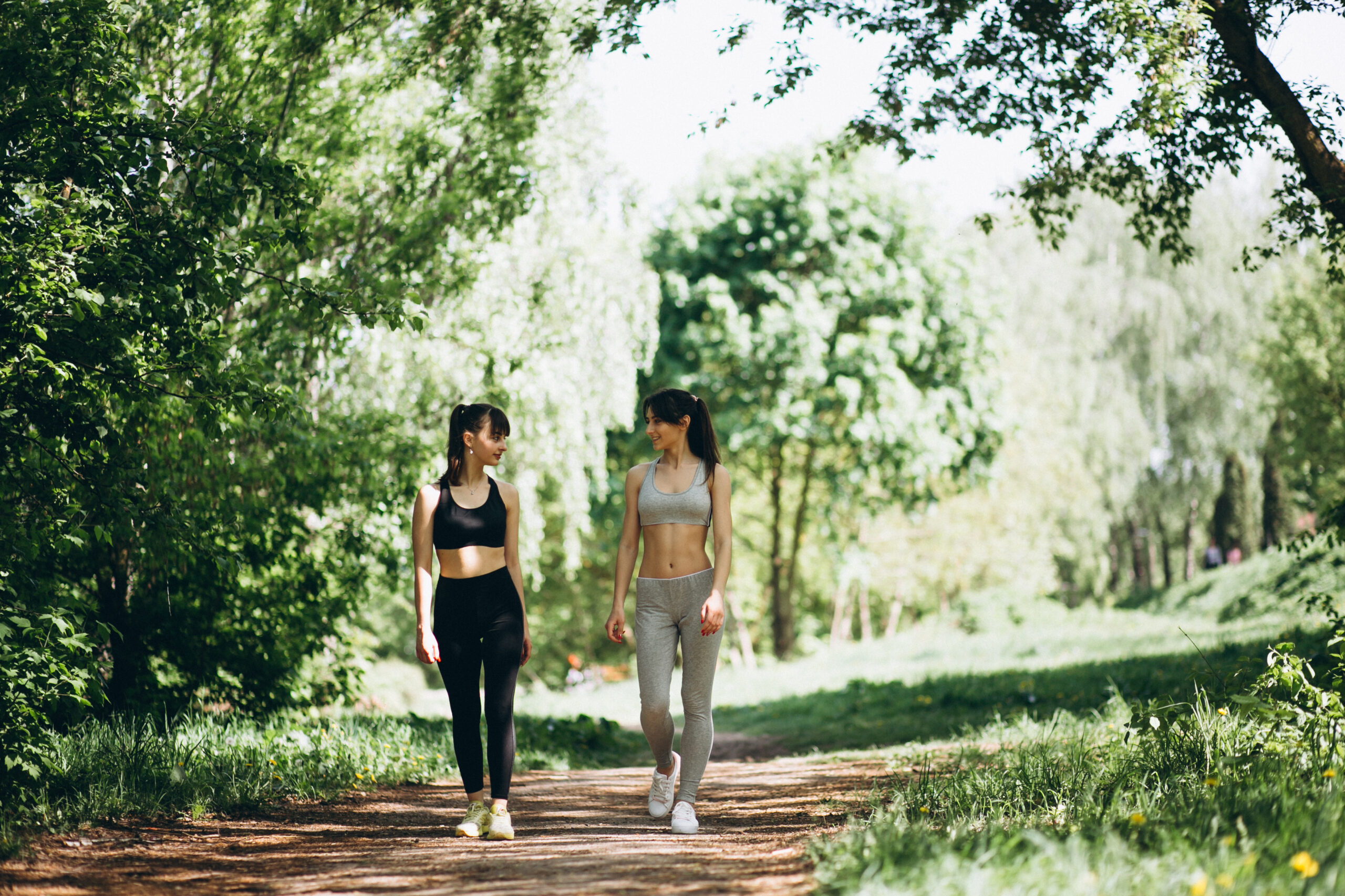 two women one in black workout clothes and one in grey, walking through a park with heads turned talking to eachother