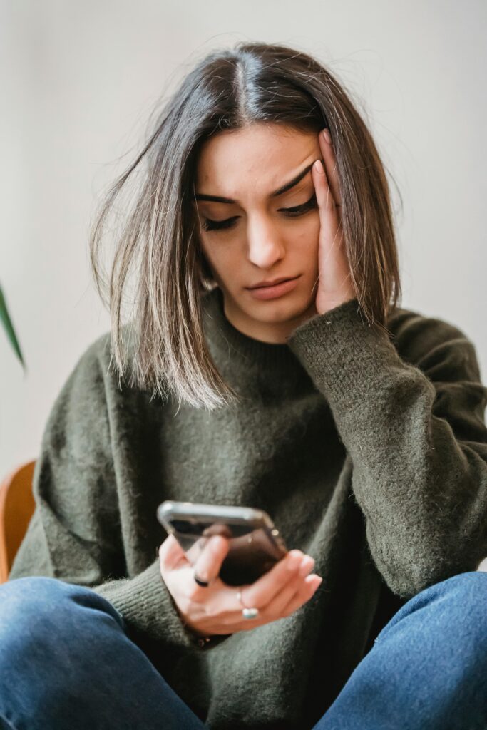woman in green jumper and jeans looking down at phone worried with hand to temple