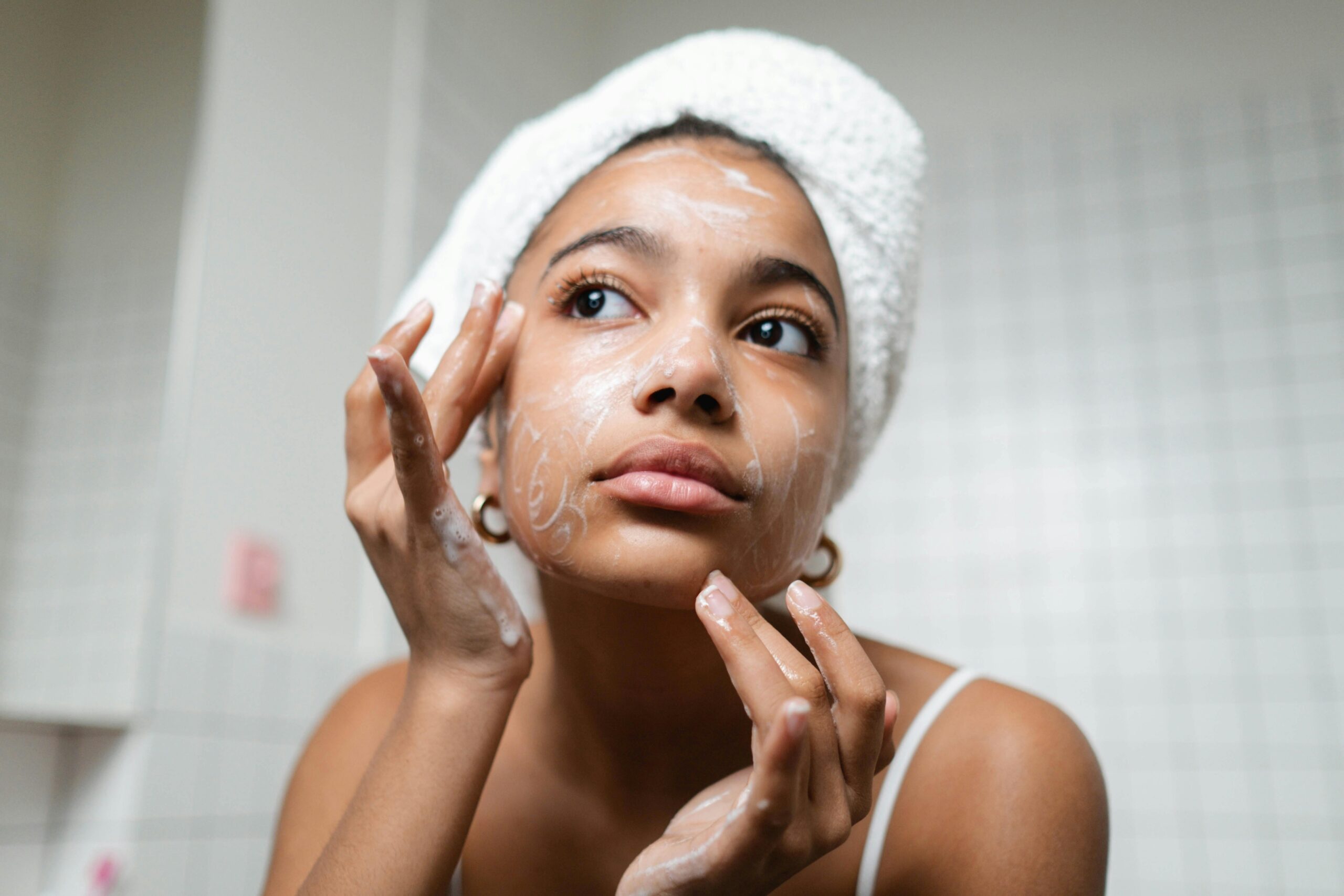 woman standing in front of a mirror face on washing face with towel on head and earrings rubbing cream wash into her face