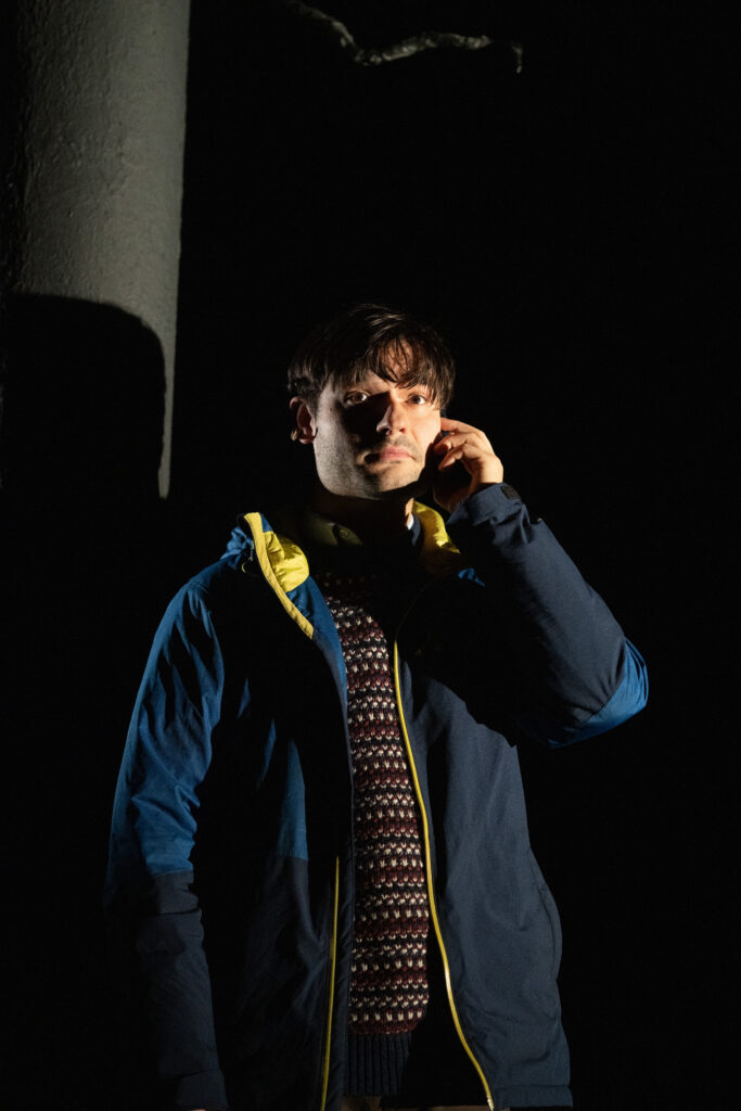 pitch black dark shadowy background with man standing in front in navy jacket with face cast in shadow