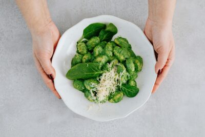 Person holding a white bowl with a green pasta dish inside
