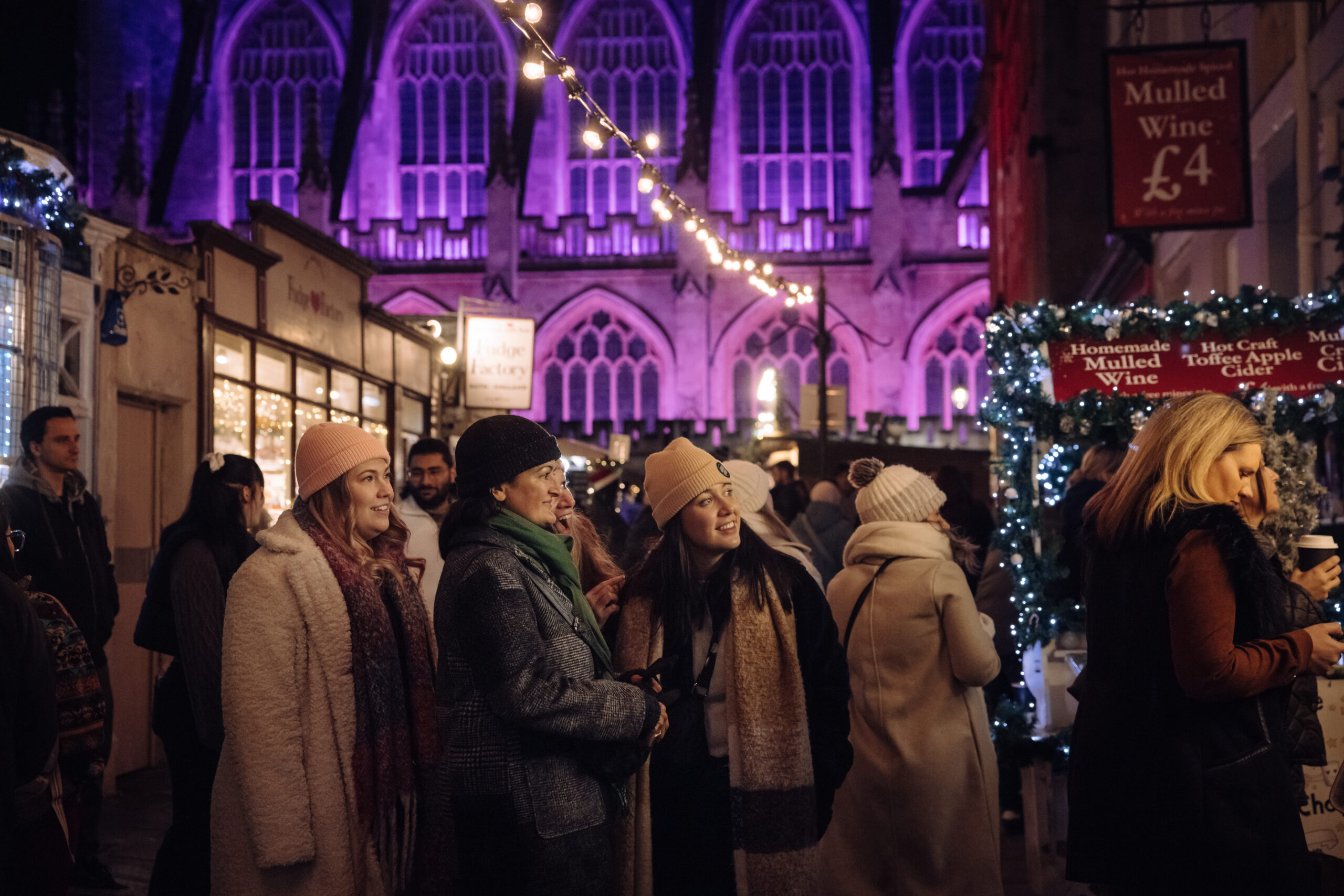 people wandering stalls at Bath Christmas market by the purple lit abbey