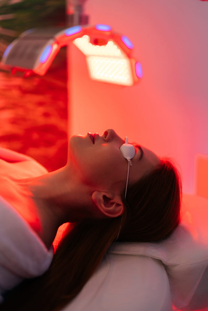 Vertical shot of young woman having red LED light facial photodynamic therapy treatment in beauty salon. Luxury medical clinic using phototherapy device with red light wavelength for acne treatments.