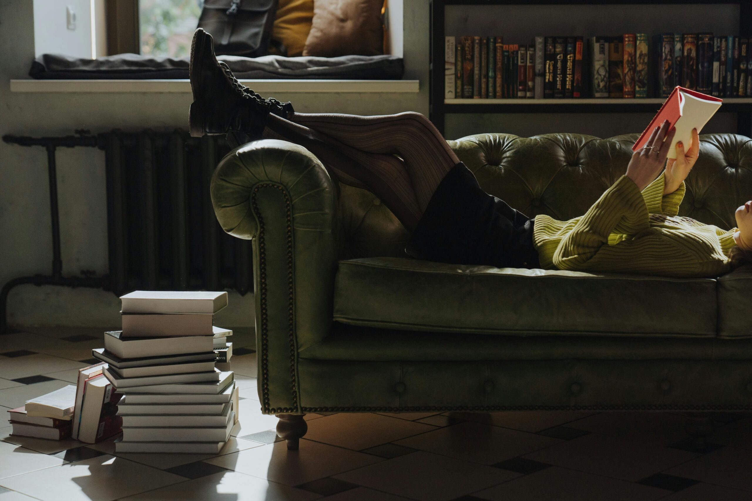 woman lying back on sofa reading a book with legs up on the arm of the sofa and a pile of books at the foot of the sofa