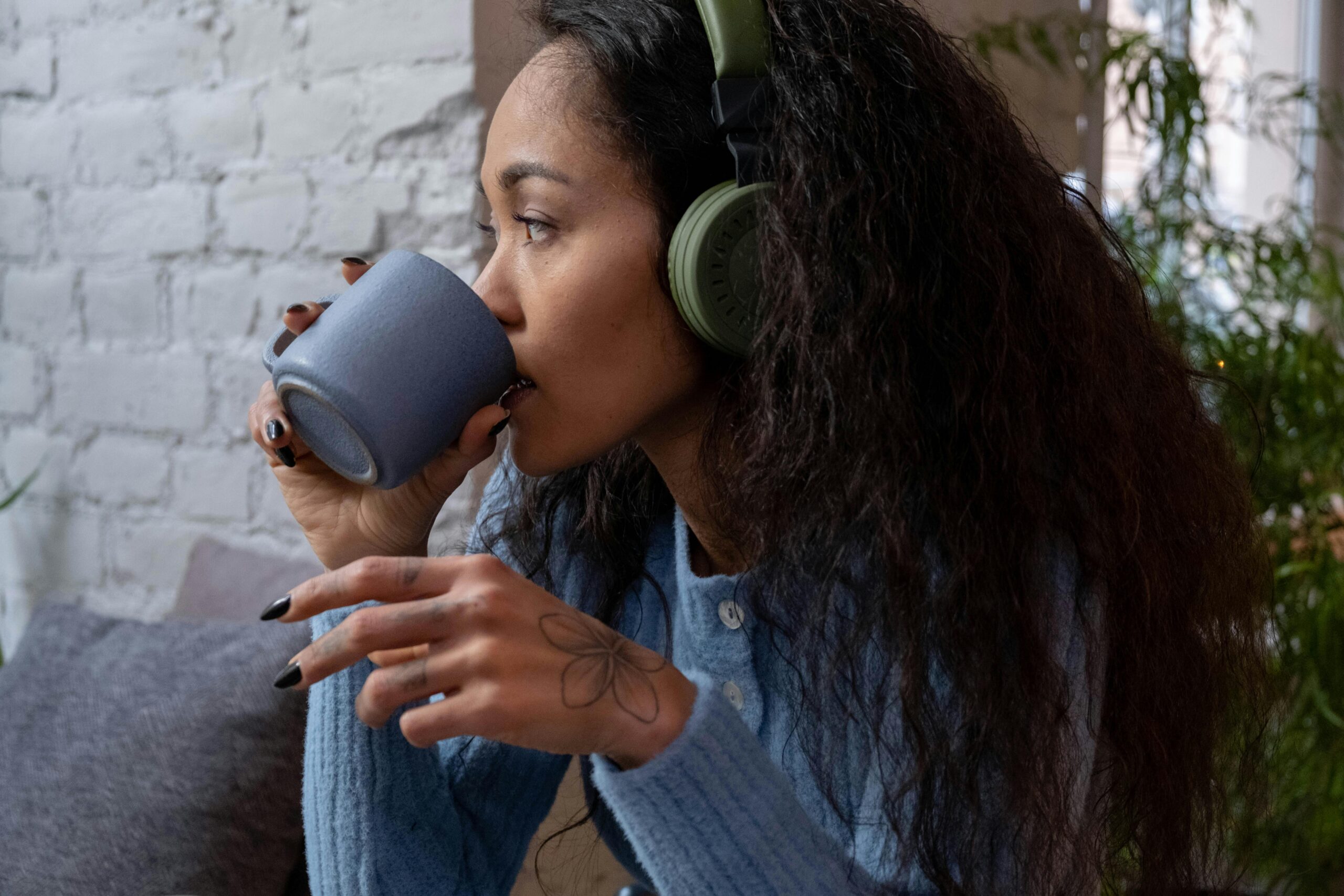 woman sits by wall in blue jumper with green headphones on sipping from a cup of coffee