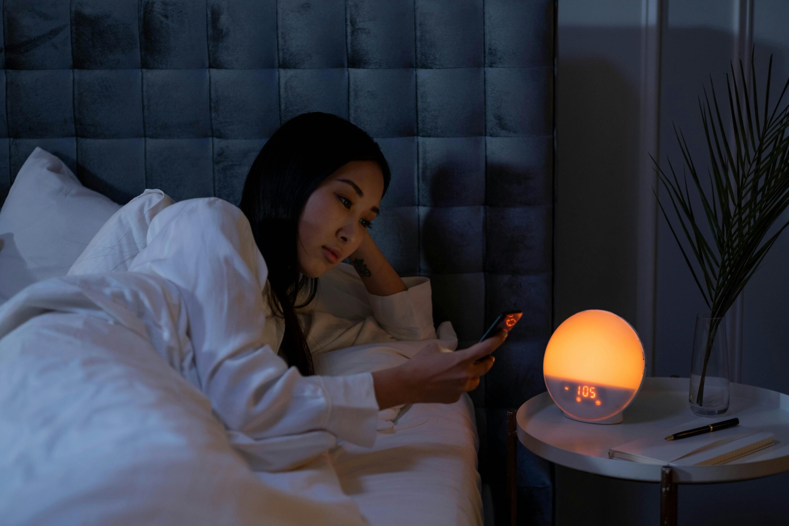 woman in bed dark room leaning over her bed to look at phone while sleep tech device alarm clock is lit up orange
