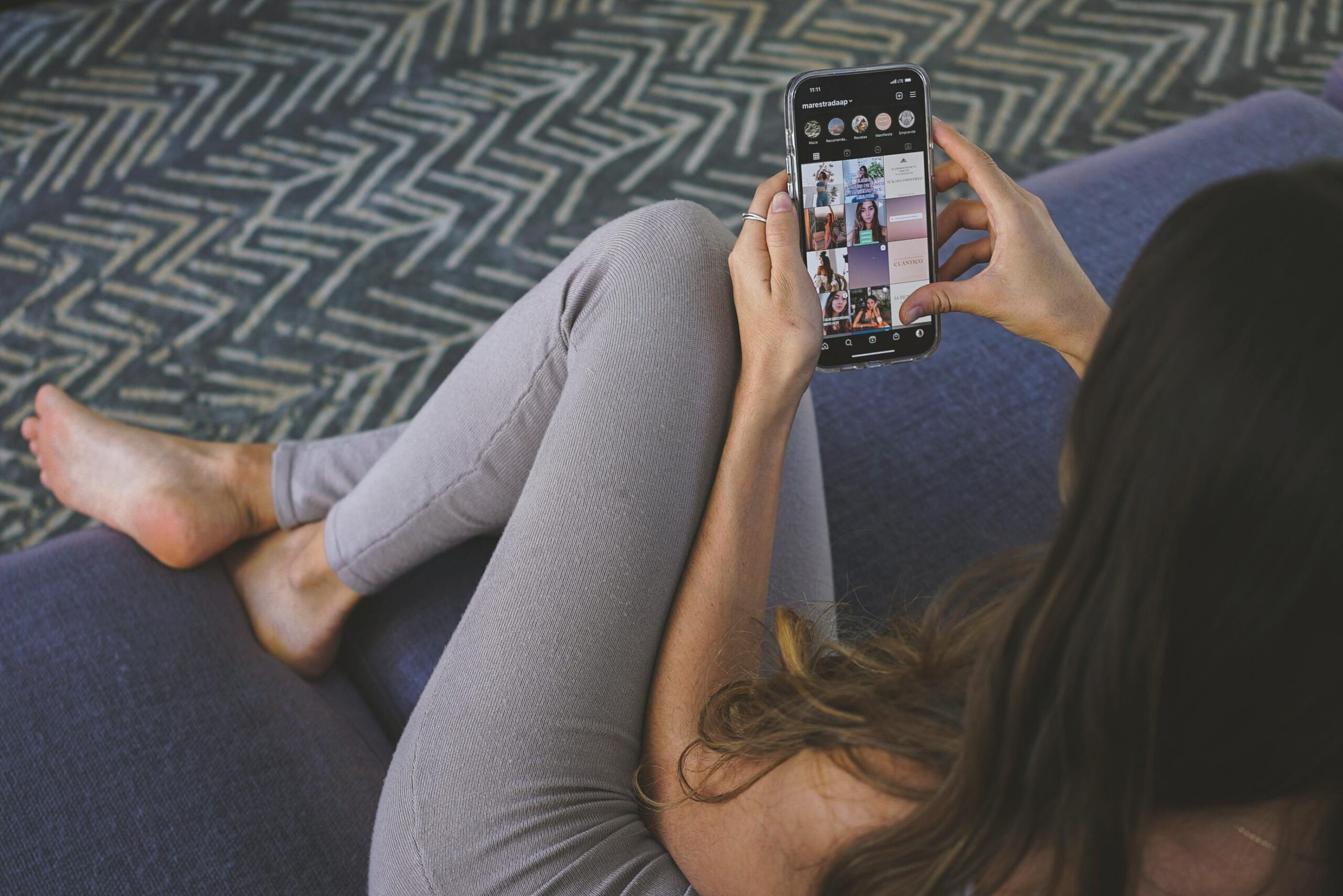 woman sitting on sofa in grey leggings scrolling through Instagram on a black iPhone