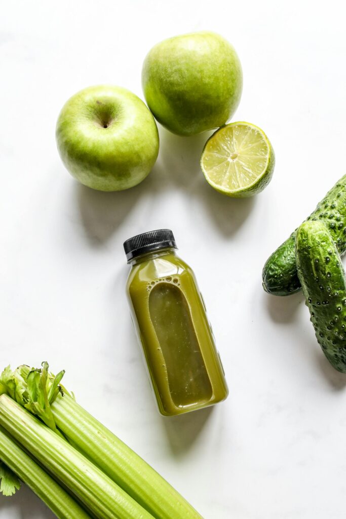 top view of marble tabletop with bottle of green juice surrounded by apples pickles and celerty