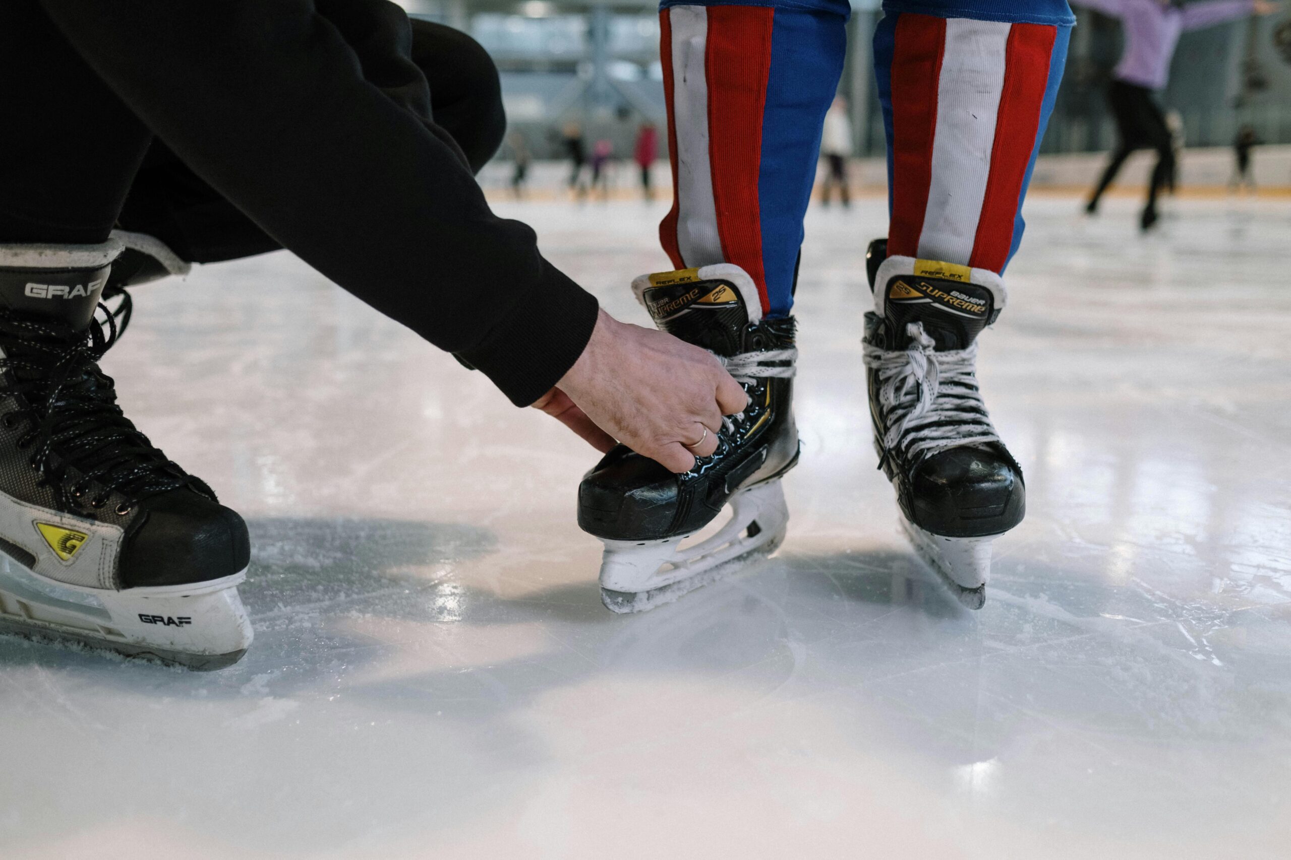 persons hands leaning down to tie skate belonging to someone else while standing on ice