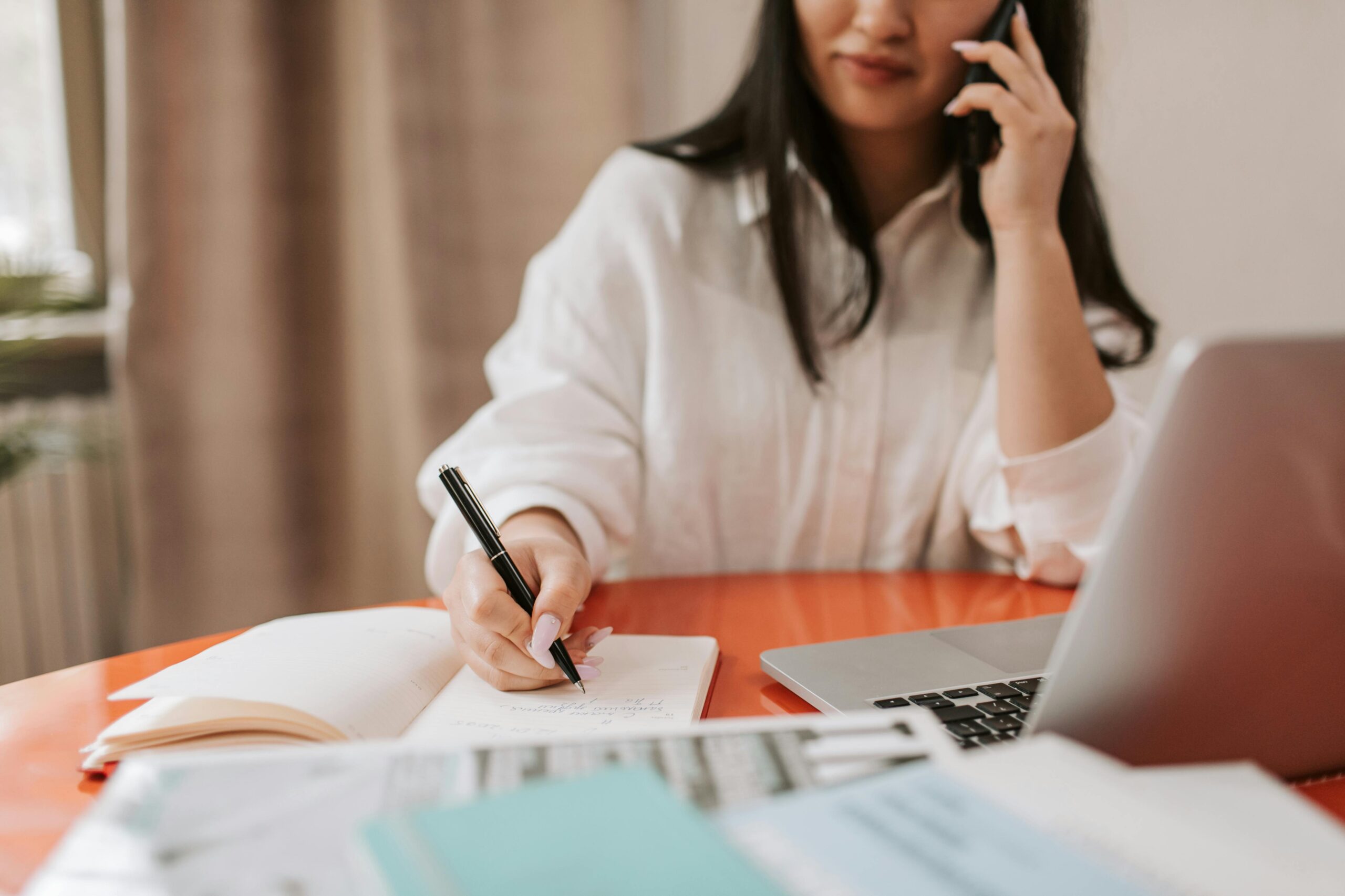woman sitting down at orange work desk writing down on a pad and on phone in front her laptop