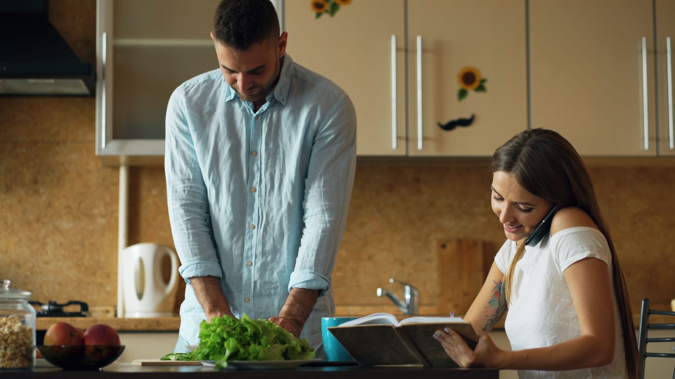 Couple at table woman looking at book on the phone while man prepares dinner