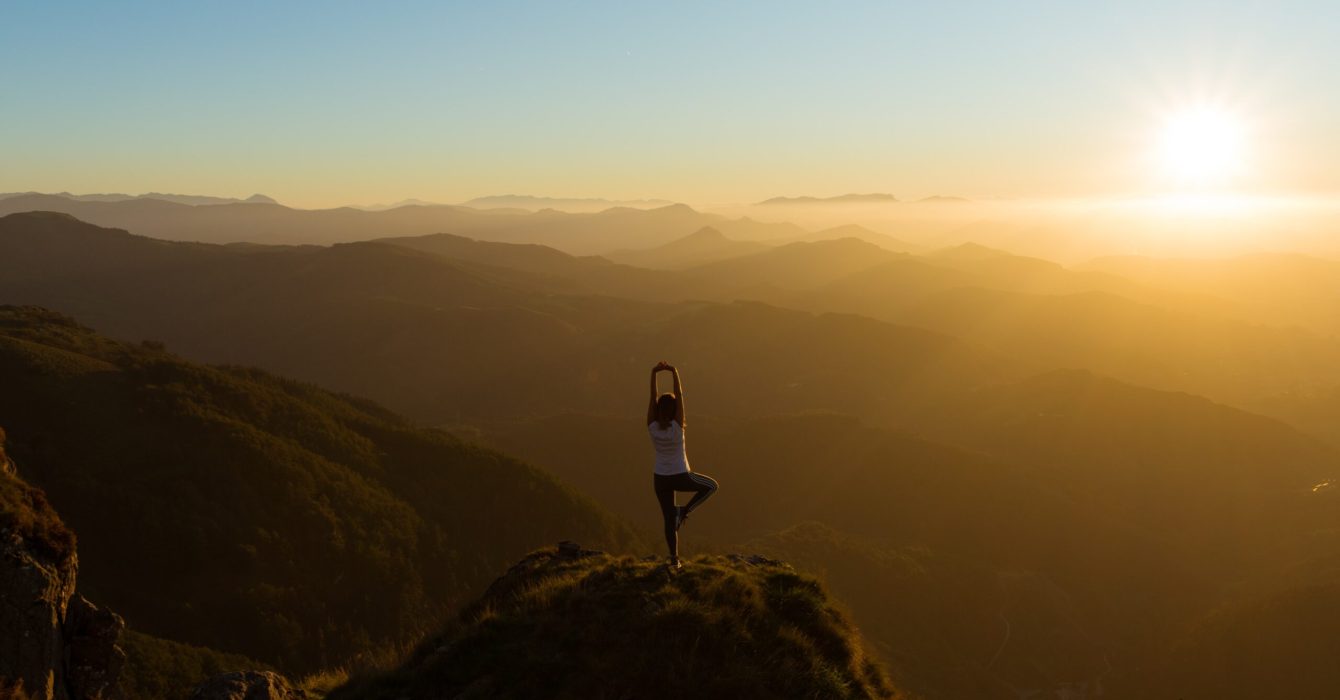 A woman practising yoga on the top of a hill as the sun sets on the horizon.