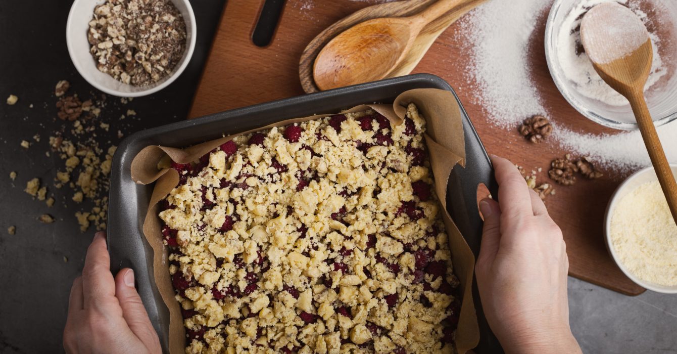 Baking cornmeal crumble bars with raspberry and walnut. Close up of hands holding tray on background of messy kitchen table, top view