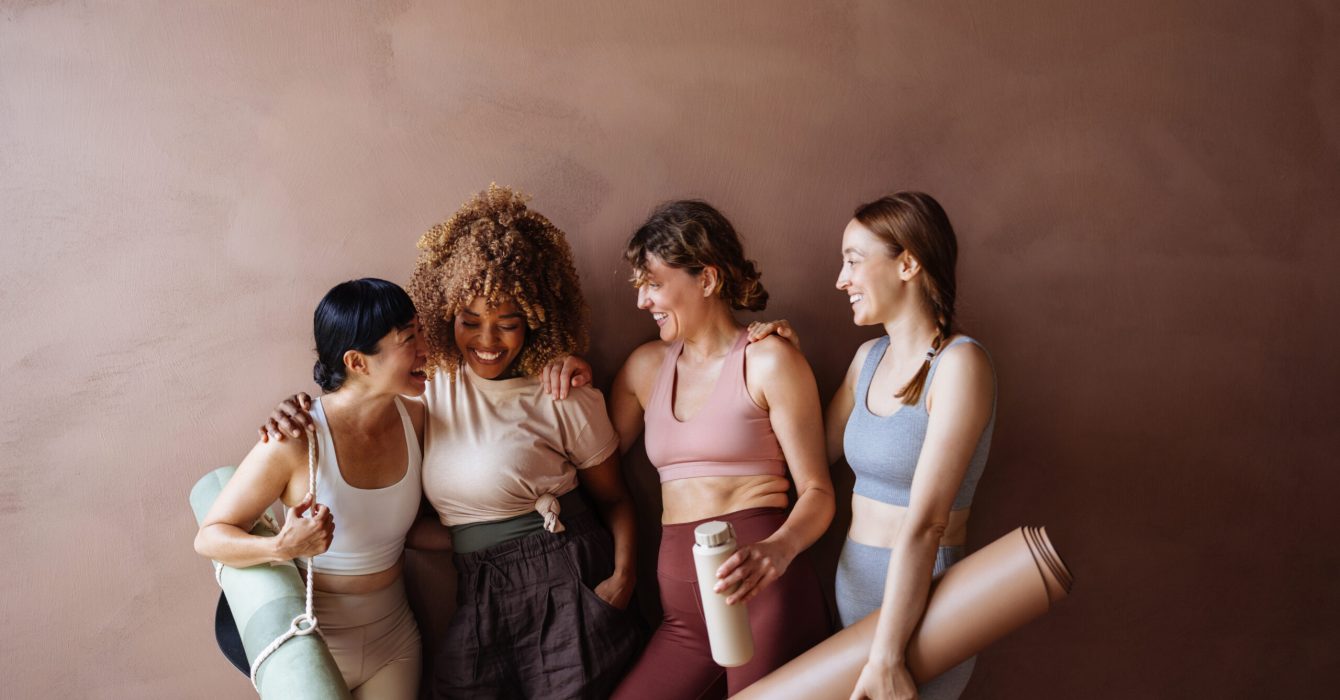 Four women in athletic outfits share a joyful moment with yoga mats and a water bottle, highlighting friendship, fitness, and wellness in a friendly studio setting.