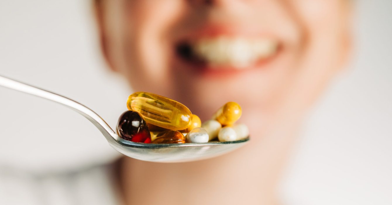 A silver spoon filled with various colorful supplements and capsules held in front of a blurred, smiling woman. This image represents proactive health management, dietary supplements, and wellness routines.