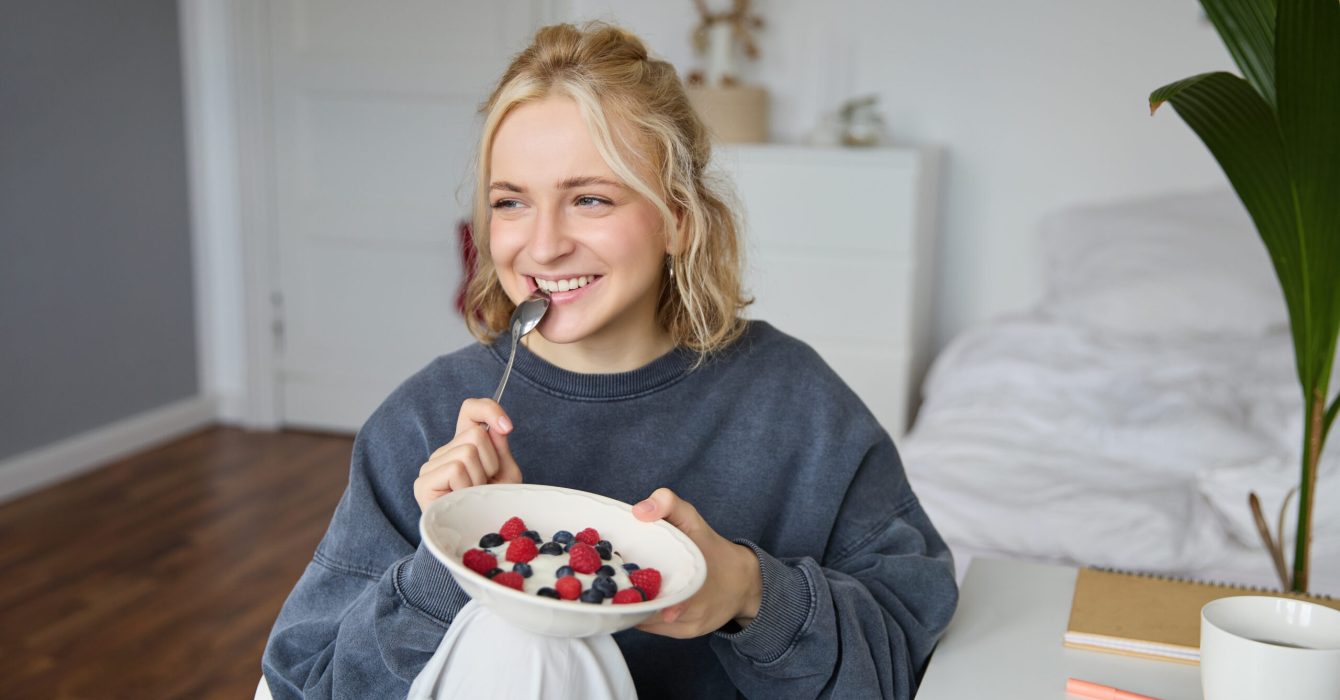 Close up portrait of smiling, cute blond woman, eating healthy lunch in her room, holding bowl with berries, biting spoon, looking happy