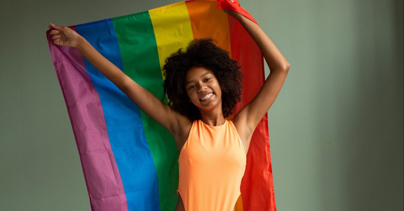 A smiling woman holds up the rainbow flag of Pride behind her.