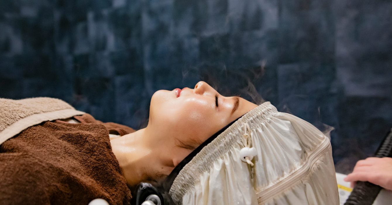 A woman lies back in a salon hair with her head being steamed