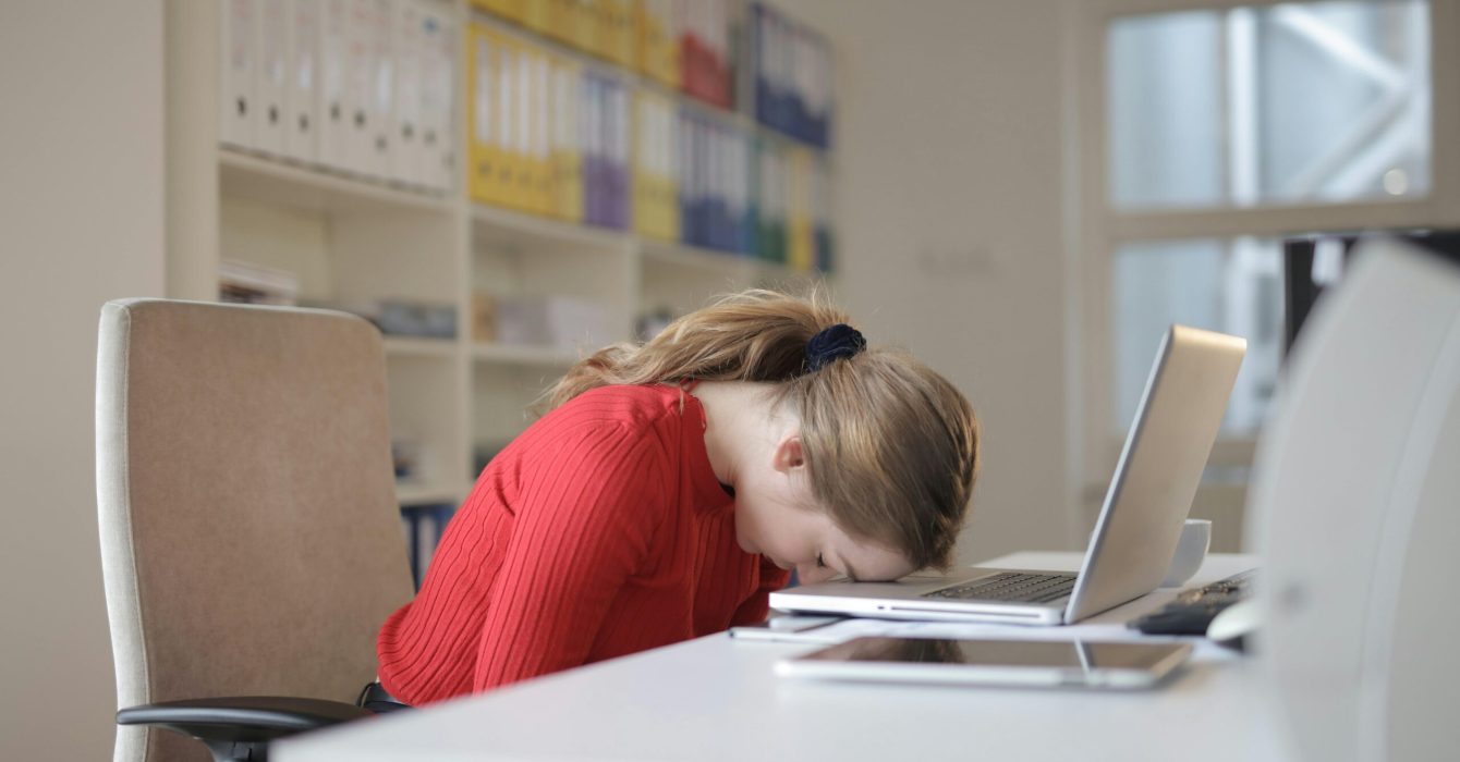 A tired woman sits at a desk with her head on the table