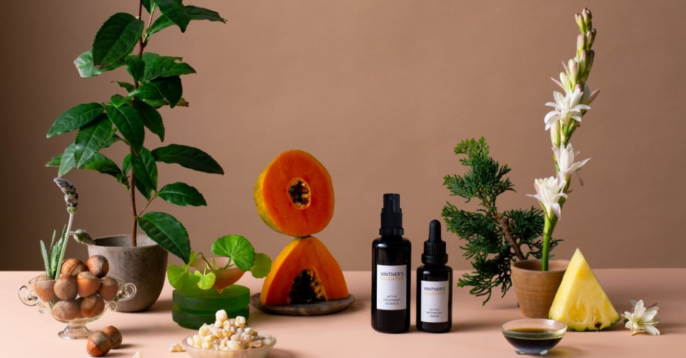 A display of two vials of Vintner’s Daughter's beauty products next to a selection of sliced fruit, green plants and bowls of nuts.