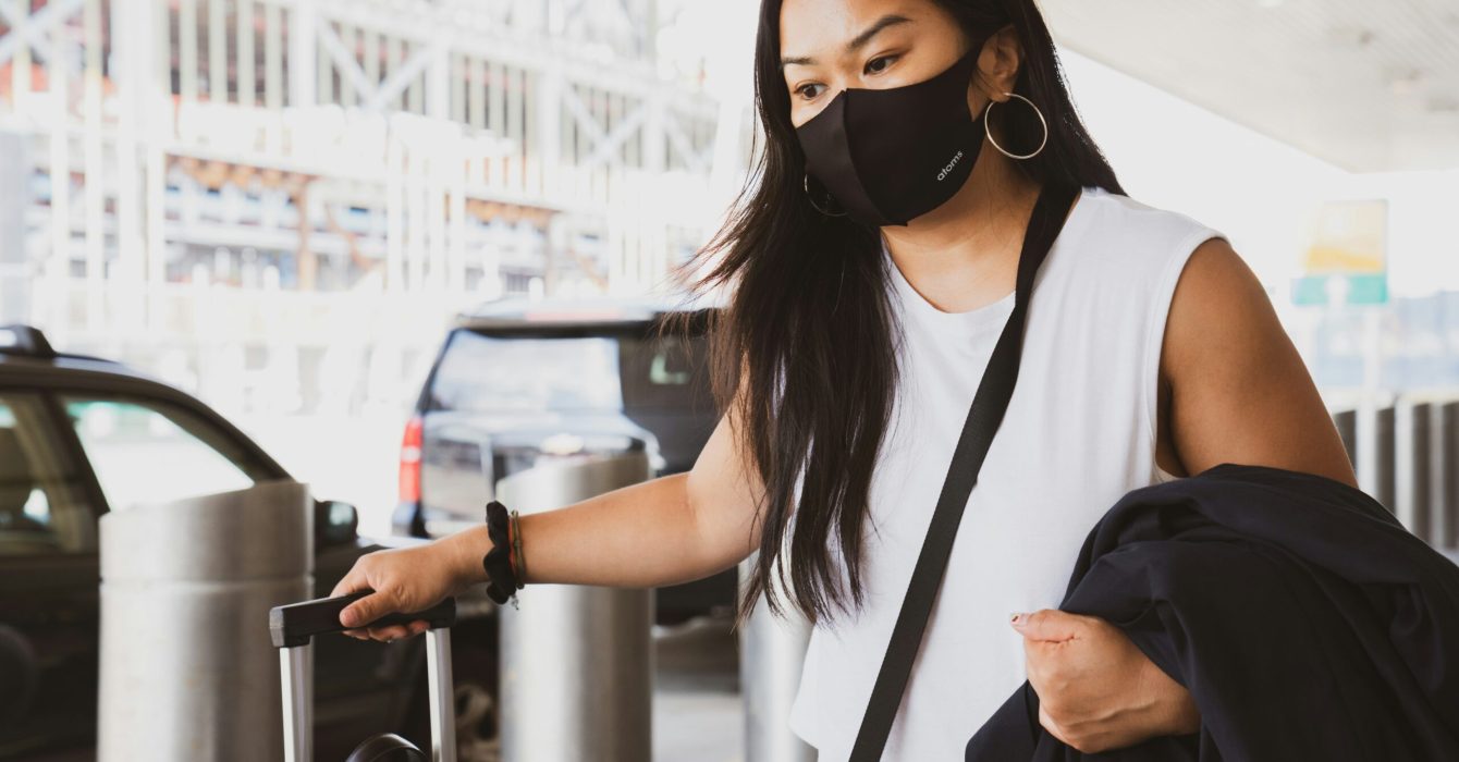 Woman outside airport wearing a face mask and pulling a suitcase