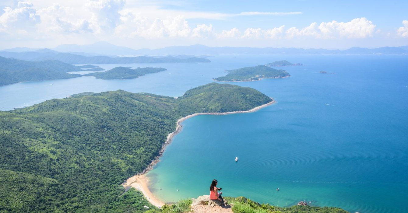 A beautiful shot of a woman sitting on a cliff with a landscape of forested hills and a blue ocean