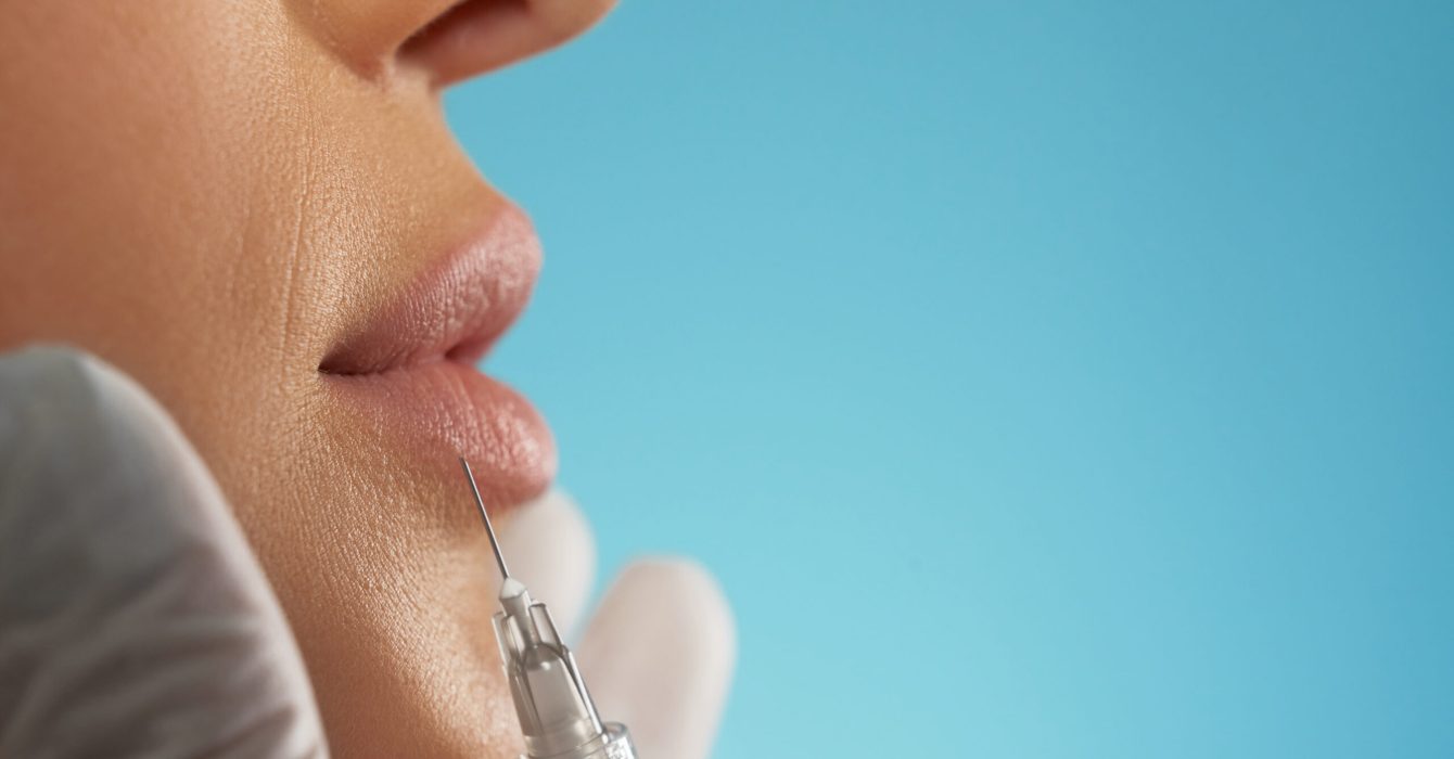 Close up of cosmetic botox injection in female lips. Side view of cosmetologist using small syringe with special liquid to make lips bigger, isolated on blue. Concept of cosmetology, beauty.