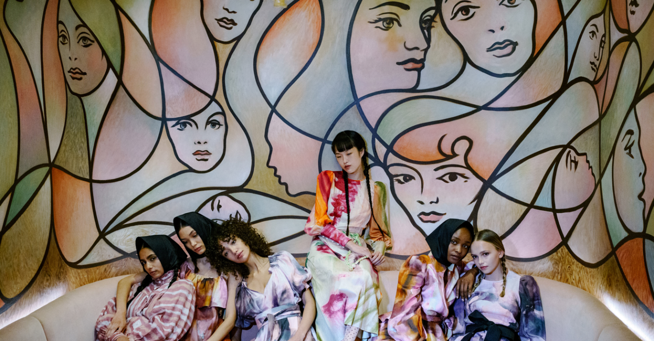 Seven international young women huddled against a mural of women's hairstyles.