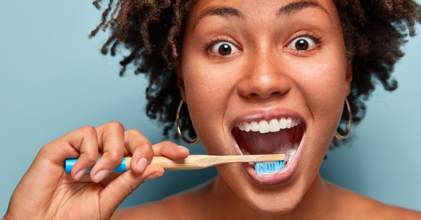 Glad mirthful woman with dark healthy skin, satisfied with new toothpaste, cleans teeth, opens mouth widely, has happy expression, curly hair, models over blue background. Close up portrait.