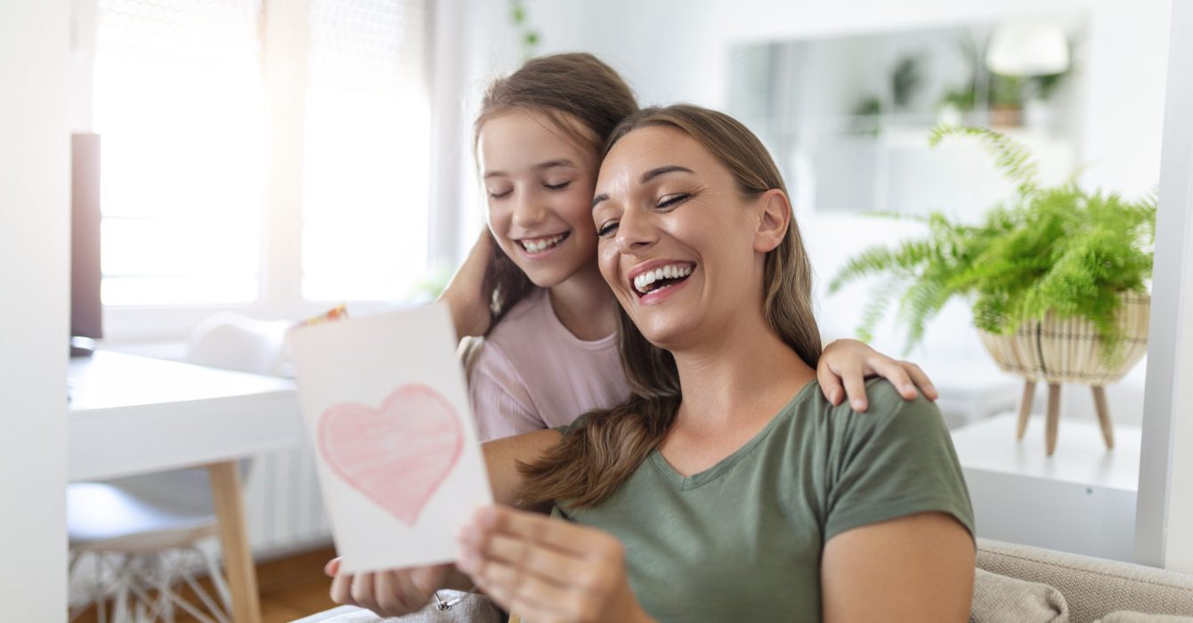 I love my you mom! Attractive young woman with little cute girl are spending time together at home, thanking for handmade card with love symbol and flowers. Happy family concept. Mother's day.