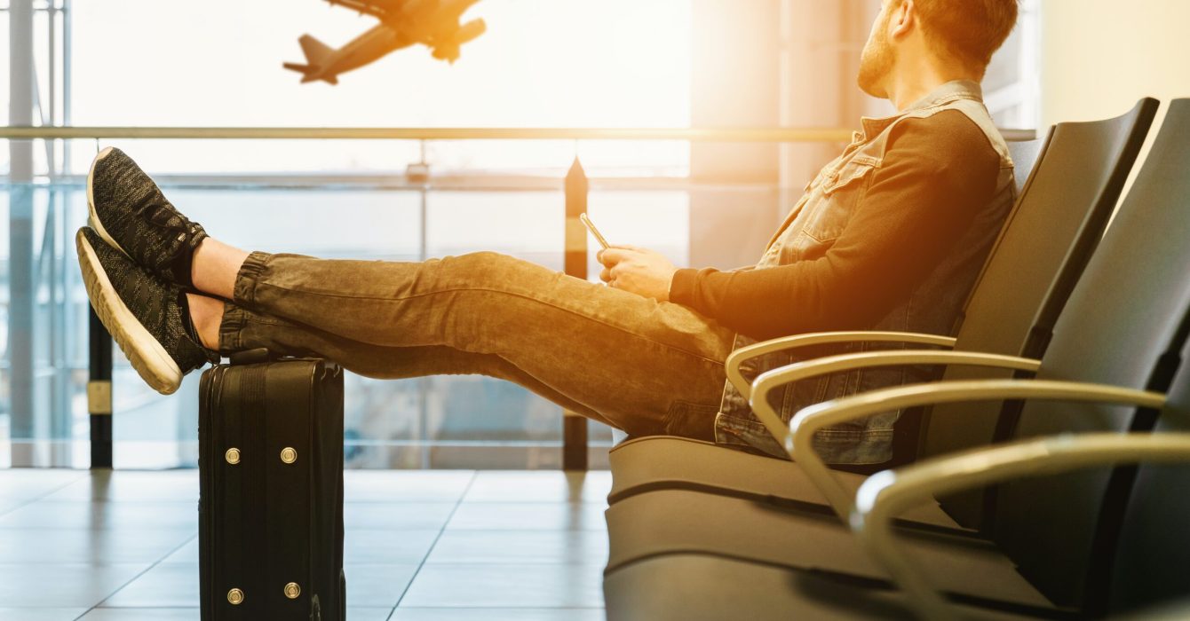 A traveller relaxes with his feet up on his suitcase in an airport lounge as a plane takes off outside the window behind him.