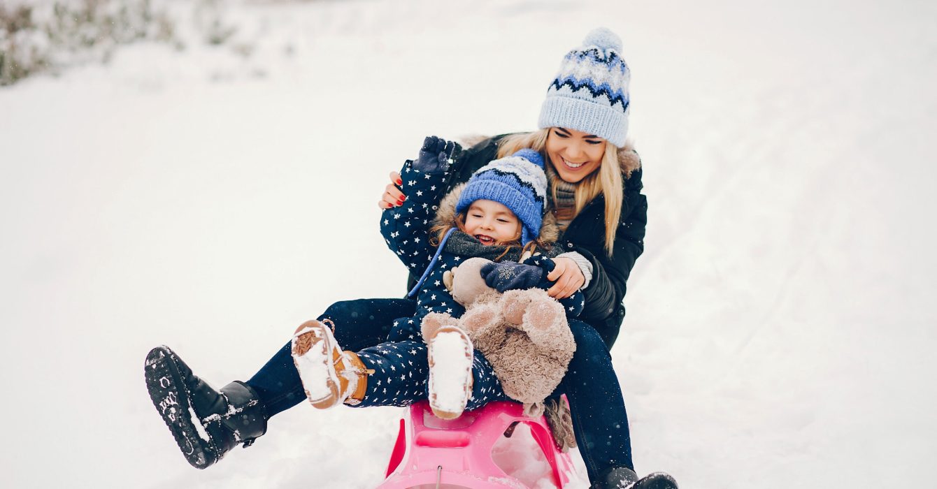 Family have fun in a winter park. Stylish mother in a black jacket. Little girl with pink sled