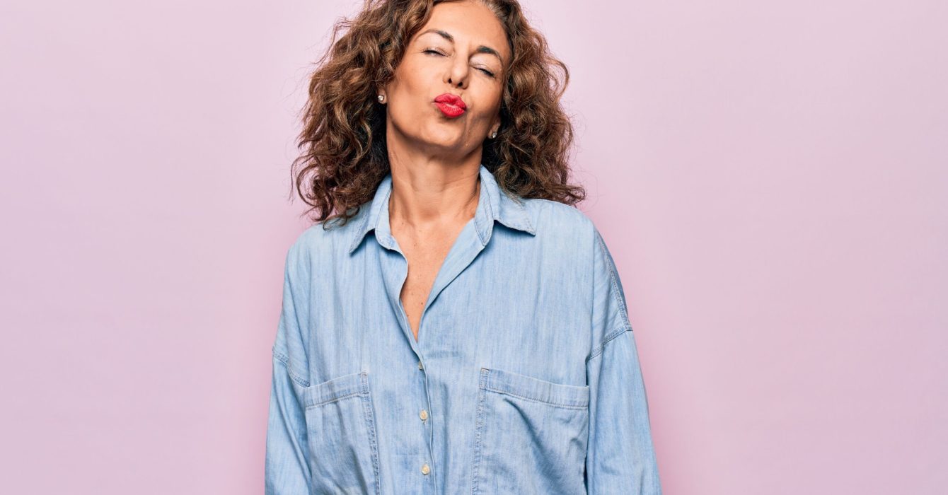 Middle age beautiful woman wearing casual denim shirt standing over pink background looking at the camera blowing a kiss on air being lovely and sexy. Love expression.