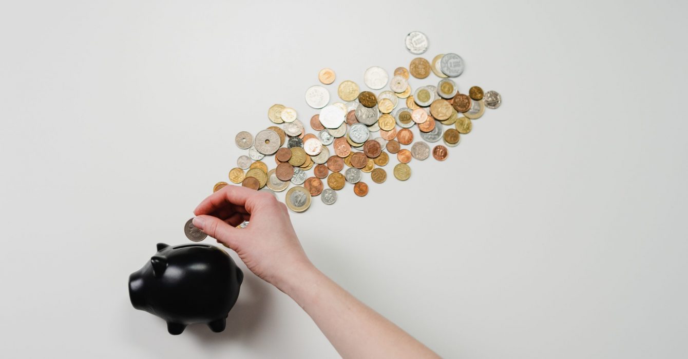 A hand placing a coin into a black piggy bank with coins streaming out from the top.