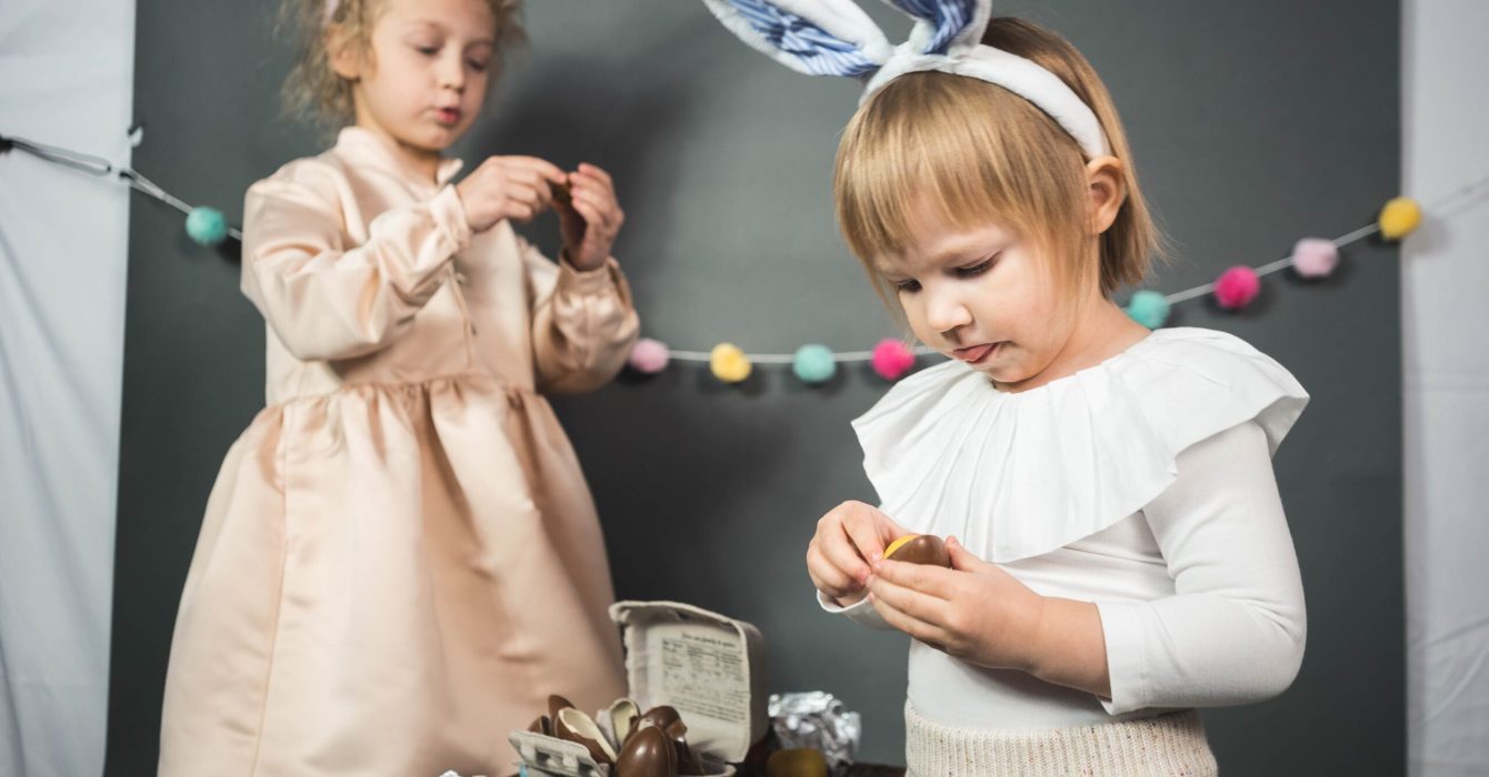 Wearing Easter bunny ears headbands, two young girls enjoy their chocolate Easter eggs.