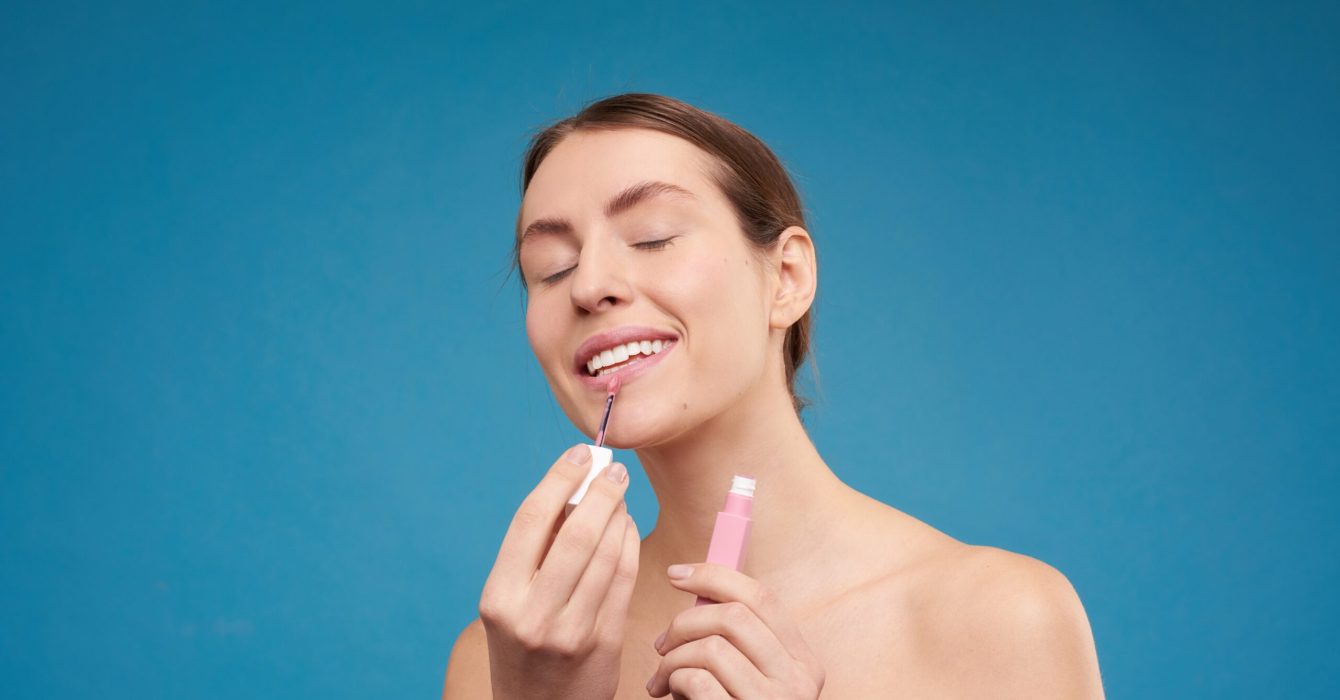 A woman applies makeup on her face against a plain blue background.