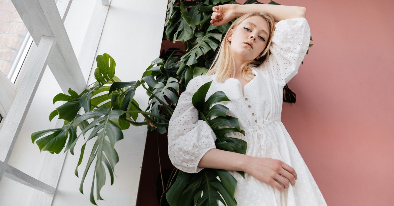A young blonde models a white summer dress against a large green plant.