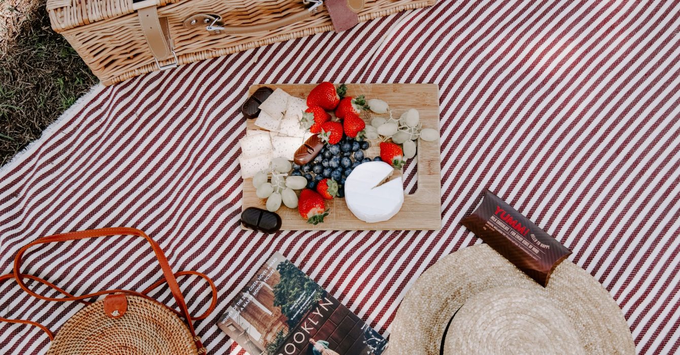 Platter with fruits and cheese, book, straw handbag and hat, and picnic basket on a striped blanket